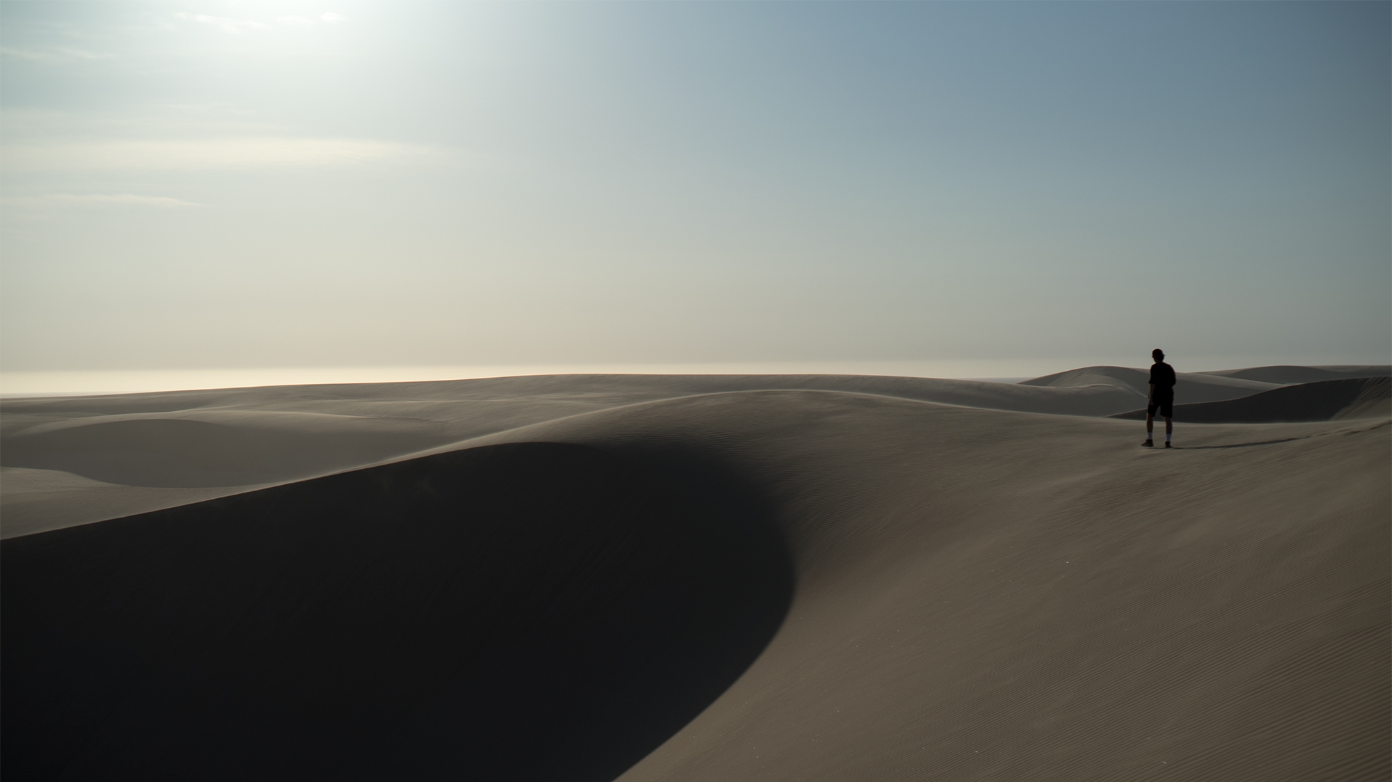 Young man walking with Bract Apparel midnight/black raglan ringer tee above large sand dune in cinematic landscape near Pismo Beach in California