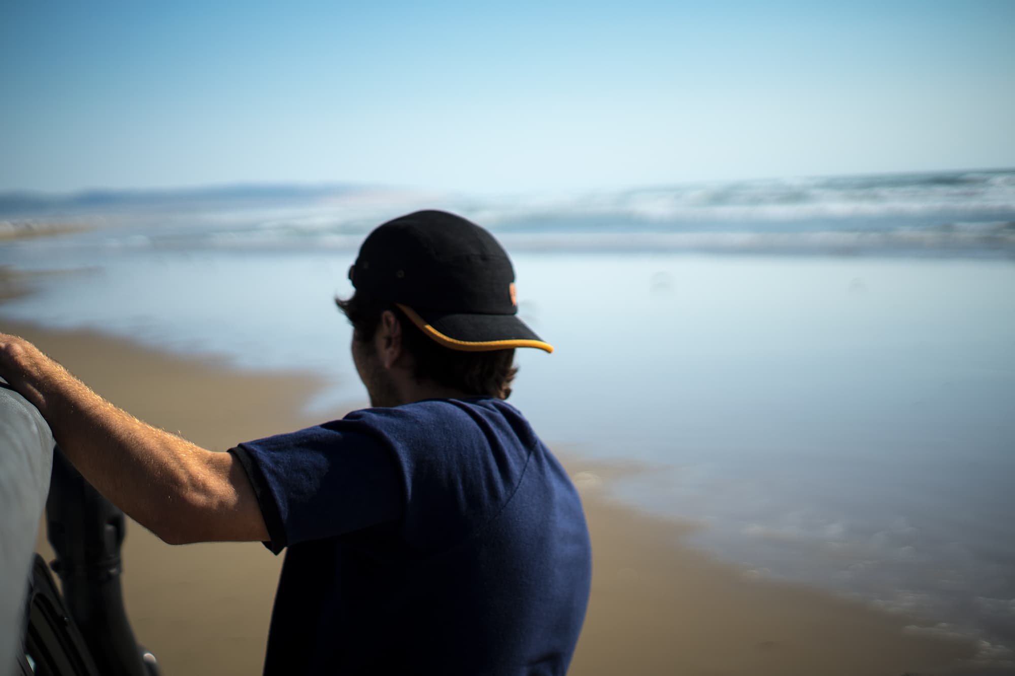 young man hanging off side of car driving on pismo beach with black bract five panel hat and midnight/black hemp and organic cotton raglan ringer tee